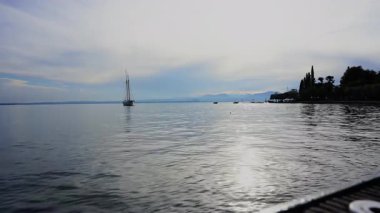 Gün batımında, İtalya, Garda Gölü 'nün sakin sularında süzülen bir yelkenlinin sakin görüntüsü. Güneşin altın ışığı su ve dağlara yansıyor..