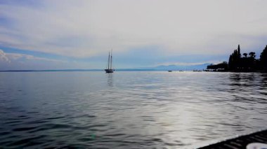 Gün batımında, İtalya, Garda Gölü 'nün sakin sularında süzülen bir yelkenlinin sakin görüntüsü. Güneşin altın ışığı su ve dağlara yansıyor..