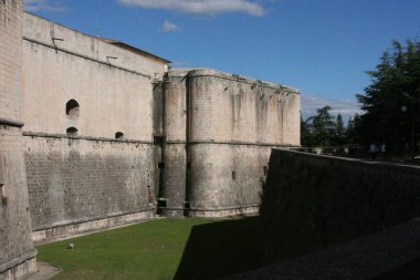 View of L'Aquila Castle with its strong, thick stone walls featuring cylindrical towers and grassy moat, against a backdrop of a bright blue sky with clouds. High quality photo clipart