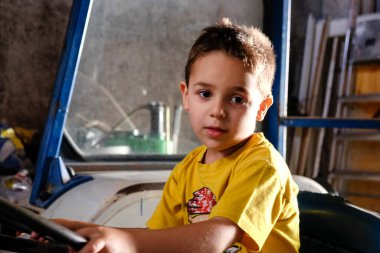 Parlak sarı tişörtlü genç bir çocuk çiftlik traktörünün direksiyonuna mutlu bir şekilde tutunuyor ve kırsal alanda oyun zamanının tadını çıkarıyor. Yüksek kalite fotoğraf
