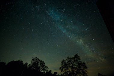 Gökyüzünde Samanyolu Galaksisinin yer aldığı bir gece manzarası, ağaçların siluetleri ve ön planda bir ev, kırsal bir gecenin güzelliğini yansıtıyor. Yüksek kalite fotoğraf