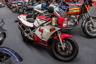 Modea, İtalya: 2025 03 Motor Festivali Fuarı Motorsiklet A kırmızı, beyaz ve siyah Yamaha RD500LC, 1980 'lerden beri aranan iki kulaçlı spor motosiklet, diğer motosikletler arasında kapalı alanda sergilenmektedir. Yüksek kalite fotoğraf