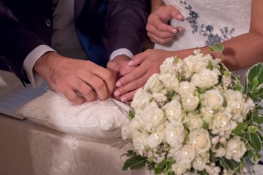 Gelin ve damadın, ön planda beyaz çiçekli bir buket bulunan beyaz işlemeli bir yastığa yerleştirilmiş altın alyanslarını değiş tokuş ederken çekilmiş yakın plan bir fotoğrafı. Yüksek kalite fotoğraf
