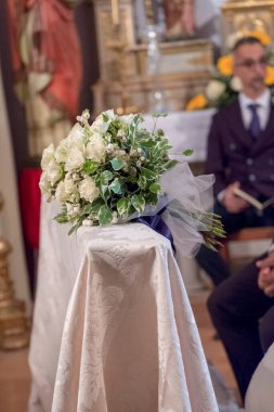Koyu mavi kurdele ve tül ile güller ve yeşillikten oluşan beyaz bir düğün buketi, arka planda bulanık figürlerle bir kilise ortamının içindeki diz çökmüş bir bankta duruyor. Yüksek kalite fotoğraf