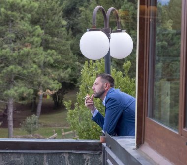 Keskin mavi takım elbiseli bir adam balkonda dikiliyor ve etrafındaki manzaraya düşünceli bir ifadeyle bakıyor. Yüksek kalite fotoğraf