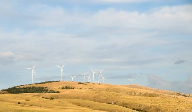 Yükselen rüzgar türbinleri, uzak bir yamaçta görkemli bir şekilde duruyor. Kılıçları, yenilenebilir enerji ve sürdürülebilirliği simgeleyen parlak bir gökyüzüne karşı dönüyor. Yüksek kalite fotoğraf