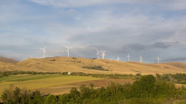 Yükselen rüzgar türbinleri, uzak bir yamaçta görkemli bir şekilde duruyor. Kılıçları, yenilenebilir enerji ve sürdürülebilirliği simgeleyen parlak bir gökyüzüne karşı dönüyor. Yüksek kalite fotoğraf
