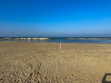 Kışın Misano Adriatico 'nun geniş, boş kumlu plajı, kumdaki lastik izleri ve açık mavi gökyüzünün altındaki uzak binalar. Yüksek kalite fotoğraf