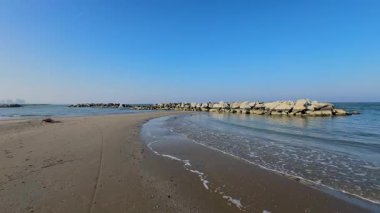 Kışın Rimini, Romagna Riviera 'da sakin bir sahilin geniş bir görüntüsü, kumlu kıyılar, sakin deniz, kayalık dalgalar ve berrak mavi bir gökyüzü, huzuru çağrıştırıyor. Yüksek kalite 4k görüntü