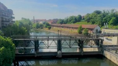 İtalya 'nın Melegnano kentindeki nehir barajı ve metal köprü yapısının manzarası, tarihi tuğla binalar ve güneşli nehir kıyıları boyunca uzanan yeşil ağaçlar. Yüksek kalite 4k görüntü