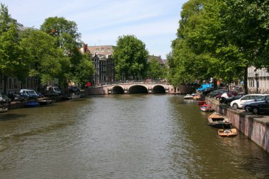 Amsterdam kanalının klasik manzarası, yemyeşil ağaçlar ve büyüleyici tarihi binalarla kaplı, arka planda kemerli bir köprü var. Yüksek kalite fotoğraf