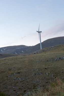 Siluetli rüzgar türbinleri, yumuşak günbatımı renkleri ve görünen ay ile dramatik bir akşam gökyüzüne karşı bir tepenin üzerinde duruyor. Yüksek kalite fotoğraf
