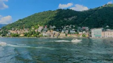 Bir sürat teknesi Como Gölü 'nün mavi sularında, yemyeşil dağlar ve güneşli bir gökyüzünün altındaki büyüleyici göl kentleri tarafından çerçevelenmiş beyaz bir uyanış yaratır. Yüksek kalite 4k görüntü