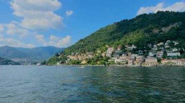 Bir sürat teknesi Como Gölü 'nün mavi sularında, yemyeşil dağlar ve güneşli bir gökyüzünün altındaki büyüleyici göl kentleri tarafından çerçevelenmiş beyaz bir uyanış yaratır. Yüksek kalite 4k görüntü