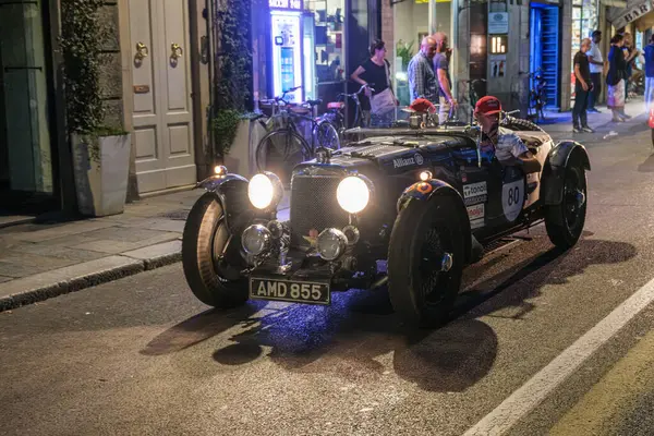 Parma, İtalya: 2025 06 20 Mille Miglia klasik rali klasik araba yarışı, parlak farları açık klasik siyah bir yarış arabası gece bir şehir sokağında insanların kaldırımda durduğu bir yolda ilerliyor. Yüksek kalite fotoğraf