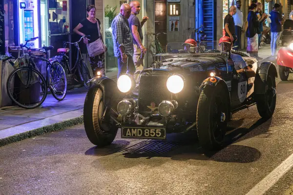 Parma, İtalya: 2025 06 20 Mille Miglia klasik rali klasik araba yarışı, parlak farları açık klasik siyah bir yarış arabası gece bir şehir sokağında insanların kaldırımda durduğu bir yolda ilerliyor. Yüksek kalite fotoğraf