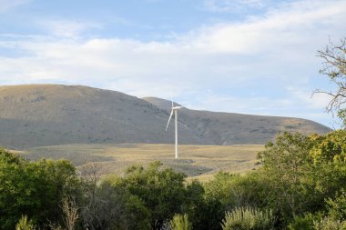 Modern bir beyaz rüzgar türbini, parçalı bulutlu bir gökyüzüne karşı yüksek bir tepede duruyor. Önünde yeşil tepeler ve ağaçlar var. Yüksek kalite fotoğraf
