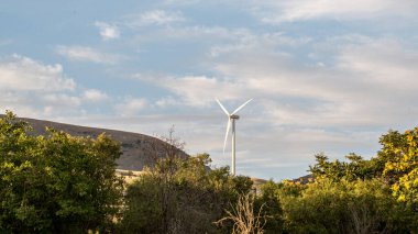 Modern bir beyaz rüzgar türbini, parçalı bulutlu bir gökyüzüne karşı yüksek bir tepede duruyor. Önünde yeşil tepeler ve ağaçlar var. Yüksek kalite fotoğraf