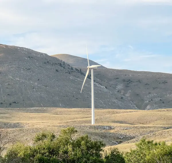 Modern bir beyaz rüzgar türbini, parçalı bulutlu bir gökyüzüne karşı yüksek bir tepede duruyor. Önünde yeşil tepeler ve ağaçlar var. Yüksek kalite fotoğraf