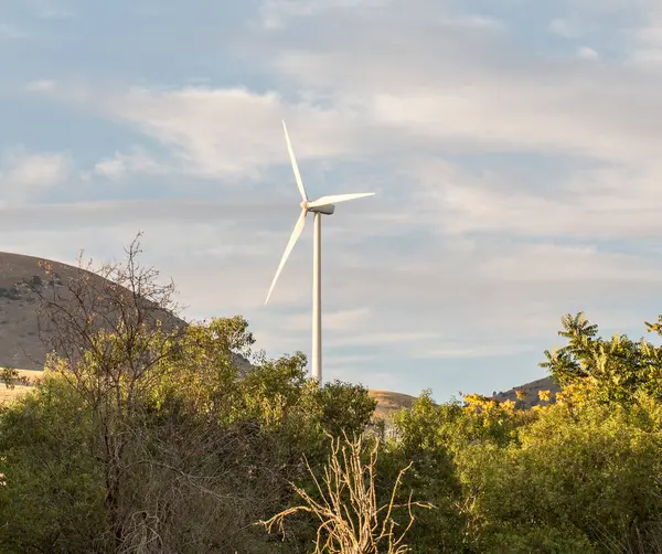 Modern bir beyaz rüzgar türbini, parçalı bulutlu bir gökyüzüne karşı yüksek bir tepede duruyor. Önünde yeşil tepeler ve ağaçlar var. Yüksek kalite fotoğraf