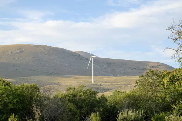 Modern bir beyaz rüzgar türbini, parçalı bulutlu bir gökyüzüne karşı yüksek bir tepede duruyor. Önünde yeşil tepeler ve ağaçlar var. Yüksek kalite fotoğraf
