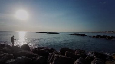 Gün batımında kıyıda kayalık bir dalgakırandan avlanan bir insanın silüeti. Güneşin sakin denize yansıması. Yüksek kalite 4k görüntü