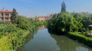 İtalya 'nın Melegnano şehrinde yemyeşil ve açık mavi gökyüzünün altındaki binalarla çevrili sakin bir nehrin manzarası..