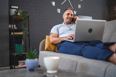 Kanepede dizüstü bilgisayar kullanırken telefonla konuşan orta yaşlı beyaz bir adam..