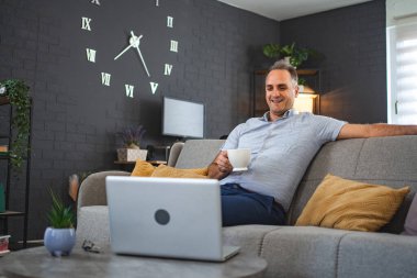 Orta yaşlı, beyaz bir adam kanepede oturup dizüstü bilgisayardan video izliyor..