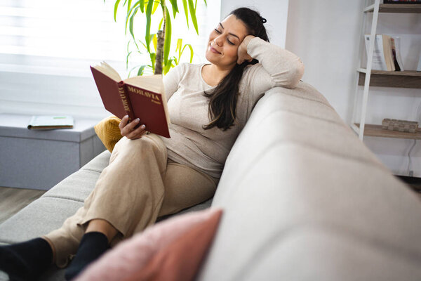 Woman reading a book while relaxing on a sofa at home.