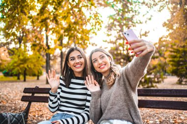 Bankta oturan iki kadın selfie çekmekle meşgul. Gülümsüyorlar ve kameraya poz veriyorlar, kollarını uzatarak tutuyorlar. Arka plan bir park ayarını gösteriyor.