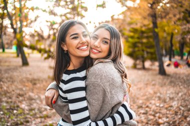 Parkta kucaklaşan iki kadın, yakınlık ve şefkat gösteriyorlar. Ağaçlarla çevrili, yeşilliklerin ortasında duruyorlar..