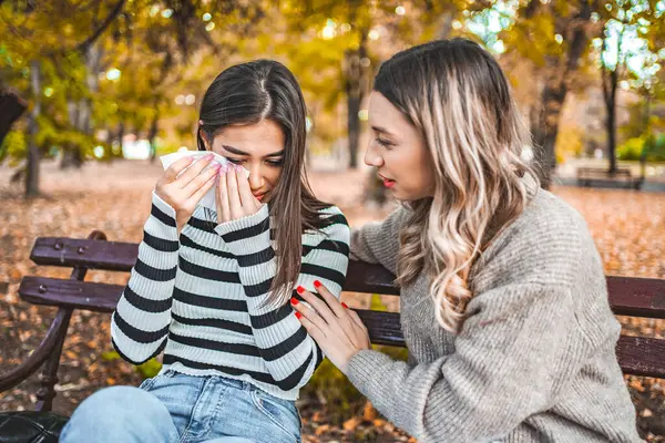 Two women are sitting on a park bench, one is comforting the other, while the other is sad and holds a handkerchief in her hand.