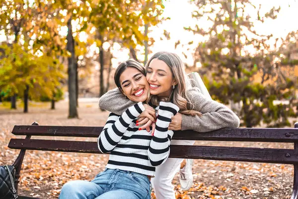 Two women of different ethnicities are kissing each other on a park bench in broad daylight. They are sitting close, leaning in towards each other, sharing an intimate moment, unaware of their