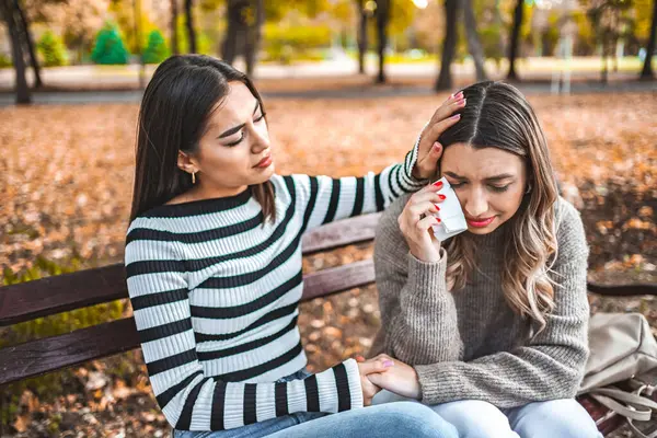 Two women are sitting on a park bench, one is comforting the other, while the other is sad and holds a handkerchief in her hand.