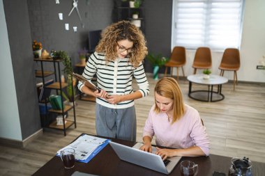İki bayan meslektaşı evden çalışıyor, biri bir masada dizüstü bilgisayar kullanıyor, diğeri ise elinde bir belgeyle birlikte bir iş raporu üzerinde çalışıyor..