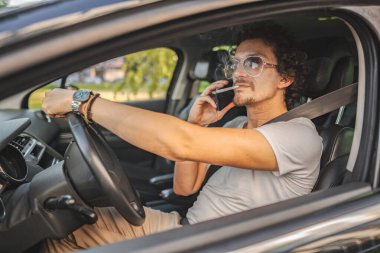 Kıvırcık saçlı ve güneş gözlüklü bir adam, araba kullanıyor, sigara içiyor ve telefon kullanıyor..