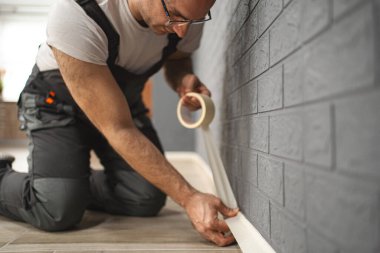 Adam duvara bant yapıştırıyor, resim yapmaya hazırlanıyor. İşine odaklanmış ve titiz görünüyor. Düz çizgiler ve doğru yerleri sağlama alıyor..