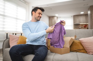 Kahverengi saçlı adam oturma odasındaki kanepede oturur ve mutlu bir şekilde bir karton paketi açar ve yeni bir tişörte bakar..