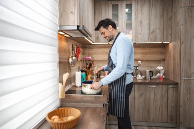 Bir adam mutfakta duruyor, tezgahta malzemeleri doğruyor ve pişiriyor. Etrafı çeşitli mutfak aletleri ve aletleriyle çevrili olarak işine odaklanmış durumda..