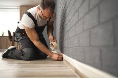 Bir adam duvara bant yapıştırıyor, resim yapmaya hazırlanıyor. İşine odaklanmış ve titiz görünüyor. Düz çizgiler ve doğru yerleri sağlama alıyor..