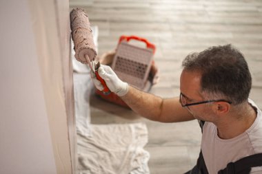 Duvarın önünde duran bir adam boyayı eşit olarak uygulamak için boya silindiri kullanıyor. Duvar sade ve yeni bir boyaya ihtiyacı var..