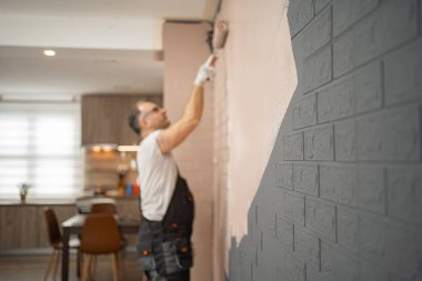 Bir adam tuğla duvarı boyayla kaplamak için boya silindiri kullanıyor. Görevine odaklanmış durumda, yüzeyde bile kapsama alanı sağlıyor..