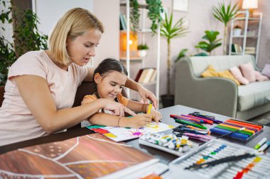 Bir anne, resim malzemeleriyle dolu bir masada renkli sanat eserleri yaparken kızına rehberlik eder..