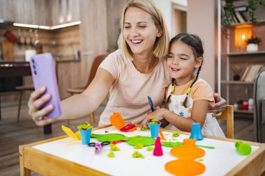 Neşeli bir anne-kız selfie için poz veriyor, masada canlı modelleme kil kreasyonlarıyla çevrili..