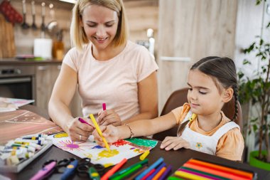 Bir anne kızının yemek masasında renkli çizimler yapmasına yardım eder. Etrafı sanat malzemeleri ve neşe dolu dekorasyonlarla çevrilidir..
