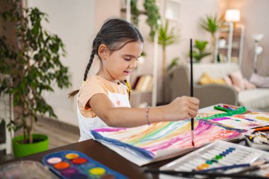 Farklı renklerde canlı resimler yapmaya odaklanmış bir kız. Kapalı bir alanda sanat malzemeleriyle çevrili..
