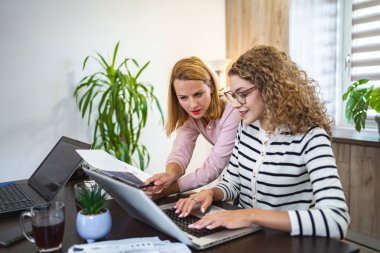 İki bayan meslektaşı evden çalışıyor, biri bir masada dizüstü bilgisayar kullanıyor, diğeri ise elinde bir belgeyle birlikte bir iş raporu üzerinde çalışıyor..
