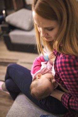 Bir anne sıcak bir oturma odasında otururken bebeğini besler..