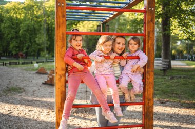 Gülümseyen ve mutlu anne, anaokulu kızı ve ikizleriyle oyun bahçesinde..